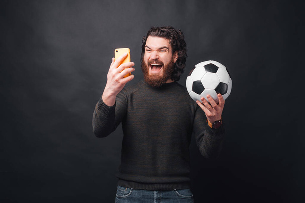 Photo of screaming amazed man looking at smartphone and holding soccer ball - Fotoğraf, Görsel