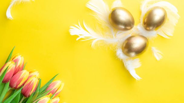 Paaseieren. Happy Easter decoratie: Gouden eieren in mand met lente tulpen, witte veren op pastelgele achtergrond. Traditionele decoratie in zonlicht. Bovenaanzicht - Foto, afbeelding