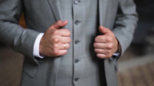 Ein kaukasischer Mann mit einem Ehering am Arm steht drinnen in einem grauen Anzug mit Weste und weißem Hemd und hält die Hände an seiner Jacke - Filmmaterial, Video
