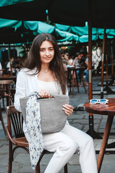 Beautiful young brunette woman wearing in sunglasses and white clothes, holding knitted bag sitting at the table of a street cafe. Copy, empty space for text. - Foto, immagini