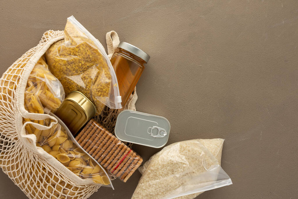 Home food supplies, necessary food for the period of quarantine and isolation, the concept of stay at home, eco bag with assorted pasta, rice and bulgur, cookies, canned food and honey,top view - Photo, Image
