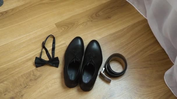 Elegantes accesorios negros de los hombres: zapatos de cordones de charol, cinturón de cuero y una mariposa se encuentran en un piso laminado de madera cerca de la ventana con tul blanco - Metraje, vídeo