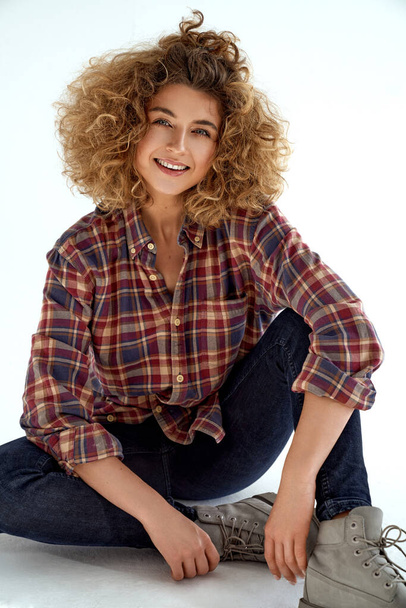 Young smiling woman with blonde curly hair wear cotton check shirt and sitting on white background - Fotografie, Obrázek