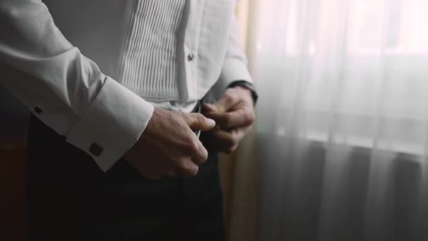 Un joven caucásico elegantemente vestido va a una reunión importante y se para junto a la ventana en el interior y se sujeta en la cintura en sus pantalones un cinturón de cuero con una hebilla de hierro - Metraje, vídeo