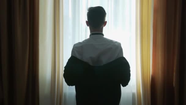 Een jonge blanke elegant gekleed man gaat naar een belangrijke vergadering en staat bij het raam binnen en trekt een jasje aan van een pak dat het over zijn schouder gooit - Video