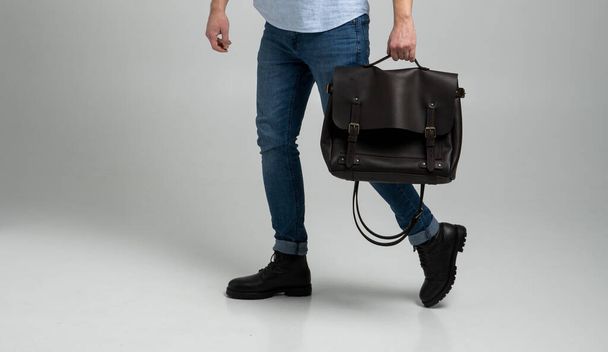 Brown mens shoulder leather bag for a documents and laptop holds by man in a blue shirt and jeans with a white background. Satchel, mens leather handmade briefcase. - Фото, изображение