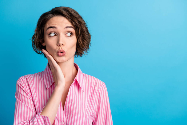 Photo of curly amazed lovely woman wear pink striped shirt look empty space arm cheekbone lips pouted isolated blue color background - Photo, Image