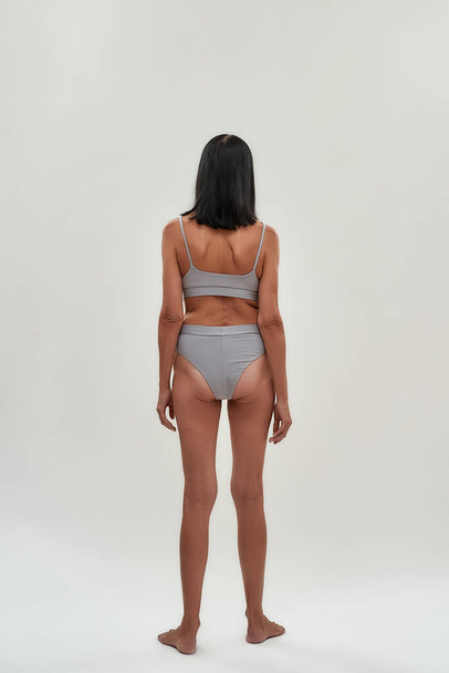 Aging process. Rear view of a half naked slim senior woman in lingerie standing against grey background in studio - Photo, image