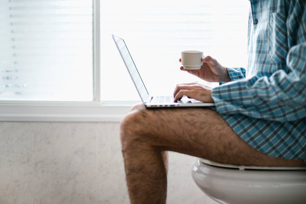 Man working on his laptop and holding a mug of coffee while sitting on the toilet at home. Telework and work from home concept. - Photo, Image