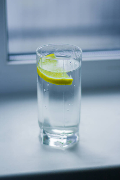 Un bicchiere d'acqua con una fetta di limone poggia su un davanzale bianco vicino alla finestra - Foto, immagini