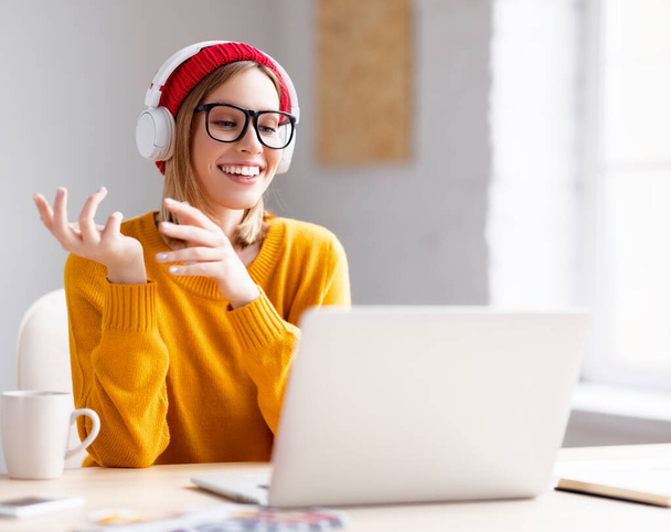 Delighted young woman in headphones and glasses smiling and gesticulating while sitting at table and talking with online colleague at home - Foto, imagen