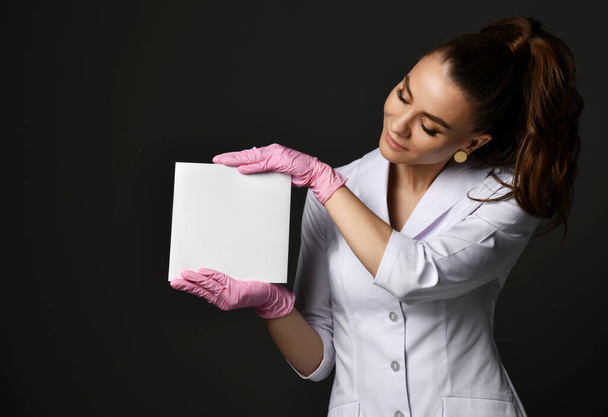 Young smiling pretty woman doctor therapist in white uniform and protective latex gloves holding blank paper sheet - Fotó, kép