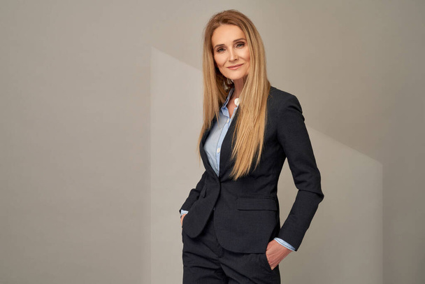 Portrait of adult woman with long blond hair posing in elegant suit over grey background in studio - Foto, imagen