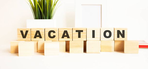 Wooden cubes with letters on a white table. The word is VACATION. White background with photo frame, house plant. - Foto, Imagem