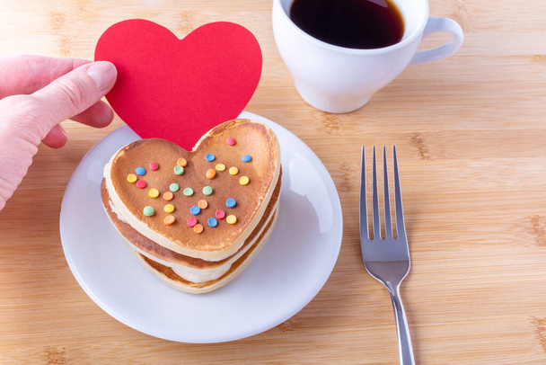 Hand holds a red blank heart shape next to homemade heart shaped pancakes with sugar decoration on a white plate, fork, mug with coffee or cocoa, close-up. Breakfast for Valentine's Day Concept - Photo, Image