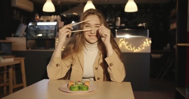 Krásná mladá dívka v béžové bundě sedí v kavárně u stolu s talířem sushi a grimasy klikání hůlky - Záběry, video