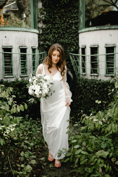 lovely young bride walks with a bouquet of flowers along the footpath in the botanical garden. floristry of a wedding bouquet. festive white flowers. Wedding Dress. beautiful modern dress. - Foto, afbeelding