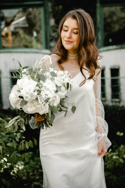lovely young bride walks with a bouquet of flowers along the footpath in the botanical garden. floristry of a wedding bouquet. festive white flowers. Wedding Dress. beautiful modern dress. - Фото, изображение