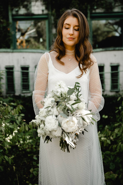lovely young bride walks with a bouquet of flowers along the footpath in the botanical garden. floristry of a wedding bouquet. festive white flowers. Wedding Dress. beautiful modern dress. - Foto, Imagem