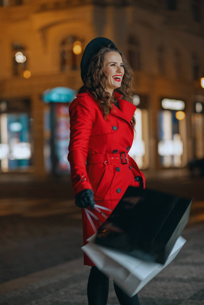 Winter Fun. happy trendy tourist woman in red coat and black beret with shopping bags outside in the city at night. - Φωτογραφία, εικόνα