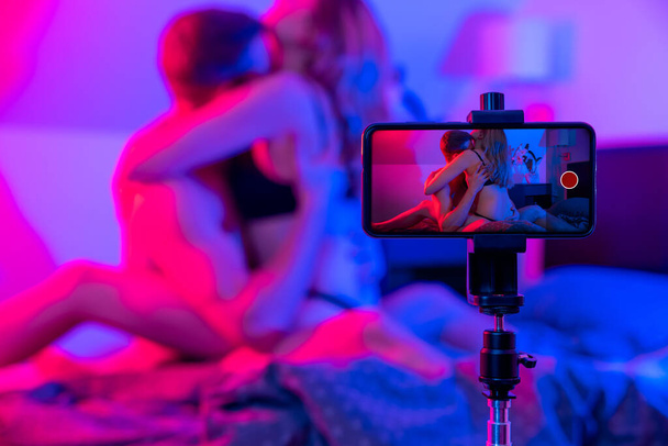 young couple in bed recording sex video with mobile phone camera. neon lights - Fotografie, Obrázek