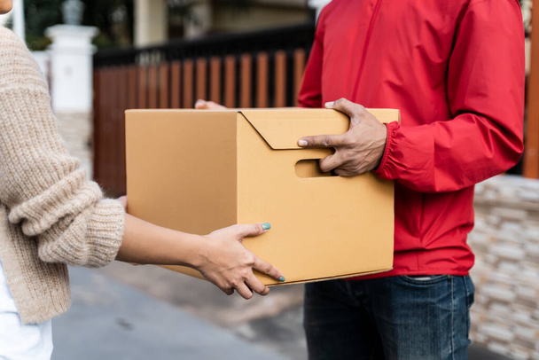 Kırmızı üniformalı Asyalı bir teslimatçı evin önünde kadın bir müşteriye paket teslim ediyor. Bir postacı ve ekspres teslimat servisi covid19 salgını sırasında paket teslim etti.. - Fotoğraf, Görsel