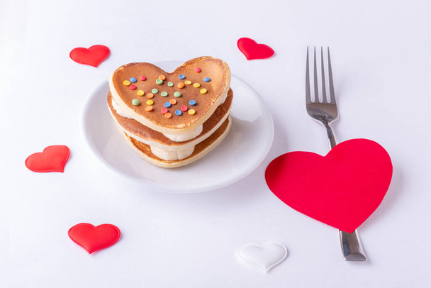 Homemade heart shaped pancakes decorated with sugar candy on a white plate, a fork and a red heart shape on a white background with hearts, top view, copy space. Breakfast for Valentine's Day - Photo, Image