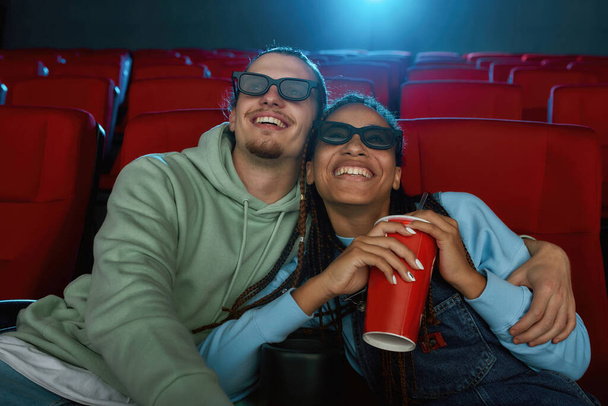 Портрет счастливой молодой пары в 3D очках, улыбающейся во время просмотра комедии вместе в кинозале и питьевой соды - Фото, изображение