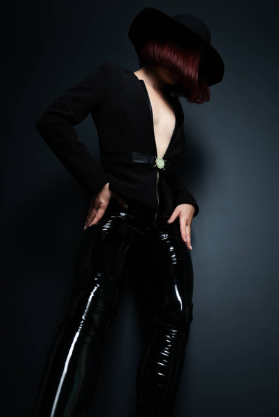 Hermosa mujer de moda con el cuerpo perfecto usando chaqueta negra, pantalones ajustados de látex y sombrero posando en el estudio sobre fondo gris profundo - Foto, imagen