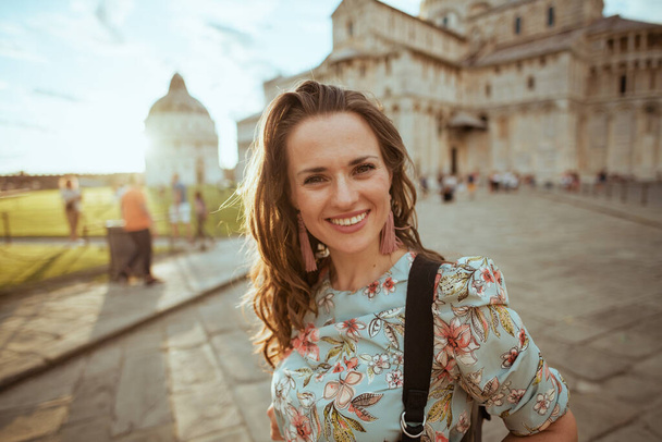 smiling stylish middle aged traveller woman in floral dress with backpack sightseeing near Duomo di Pisa. - Fotó, kép