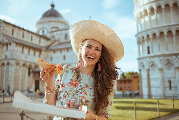 mosolygós elegáns nő virágos ruhában pizza és kalap közelében ferde torony Pisa, Olaszország. - Fotó, kép