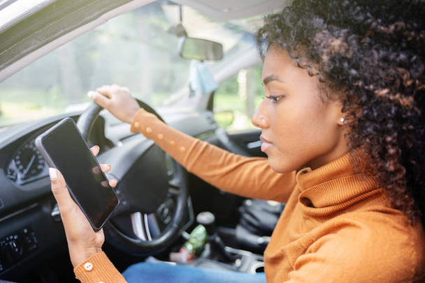Νεαρή γυναίκα οδηγεί αυτοκίνητο και ελέγχει το τηλέφωνό της. - Φωτογραφία, εικόνα