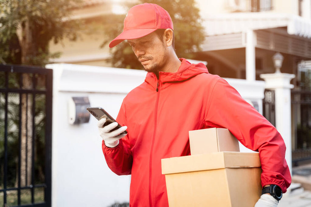 Gesneden foto van een bezorger in rood uniform houdt postpakketstands op straat kijkt naar mobiele telefoon controleren GPS-adres om dingen te leveren aan de klant. Leveringsservice tijdens COVID-19 pandemie. - Foto, afbeelding