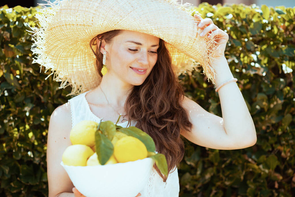 happy stylish 40 years old housewife in white shirt with plate of local farm lemons and hat outdoors near green wall. - Photo, image