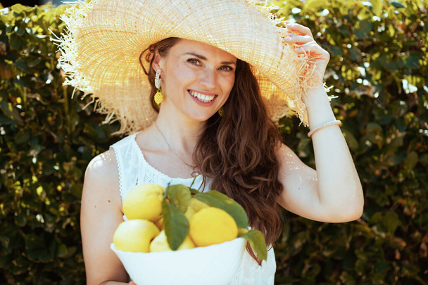 Portrait of smiling modern 40 years old housewife in white shirt with plate of local farm lemons and hat outdoors near green wall. - Foto, Bild