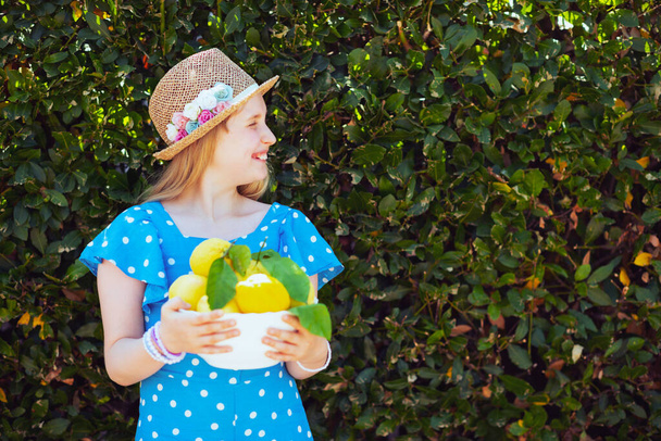 happy trendy child in blue overall and hat with plate of local farm lemons outdoors near green wall. - Valokuva, kuva