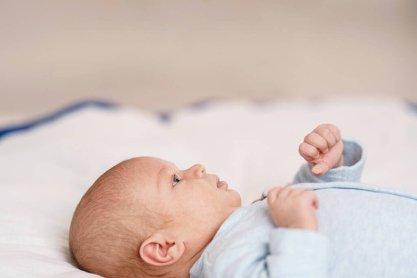Cute 2-month-old blue-eyed Caucasian baby in a blue bodysuit is lying on her back on the bed, playing with her hands and looking up. Close-up. Copy space - Fotoğraf, Görsel
