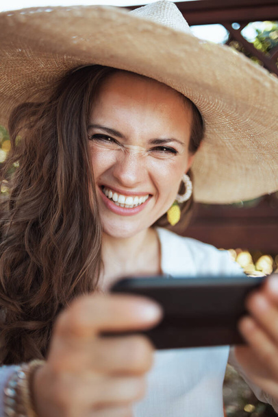 Portrait of smiling modern middle aged woman in white shirt with hat using smartphone app in the patio. - Fotoğraf, Görsel
