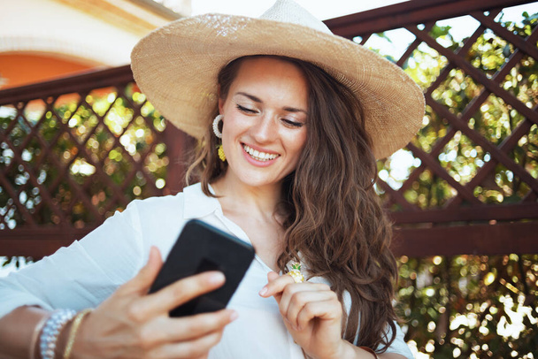 smiling young woman in white shirt with hat having video meeting on a smartphone in the terrace of guest house hotel. - Foto, immagini