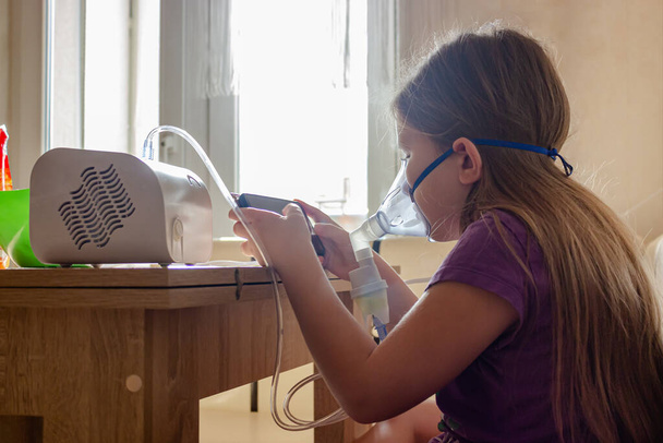 Une petite fille dans un masque fait des injections à domicile et joue à un jeu mobile afin de ne pas s'ennuyer. L'utilisation d'un nébuliseur de compresseur à la maison. - Photo, image