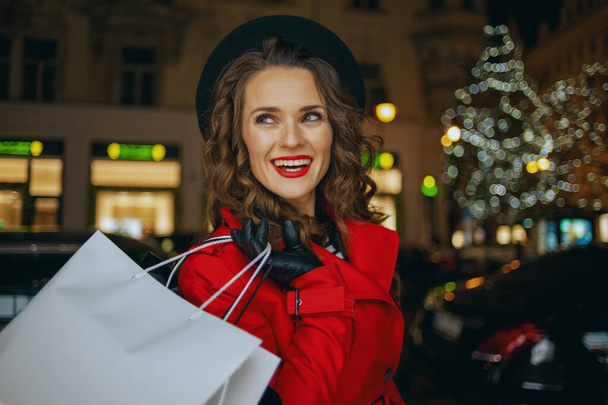 Winter Fun. smiling stylish woman in red coat and black beret with shopping bags outdoors in the city at night. - Foto, Bild