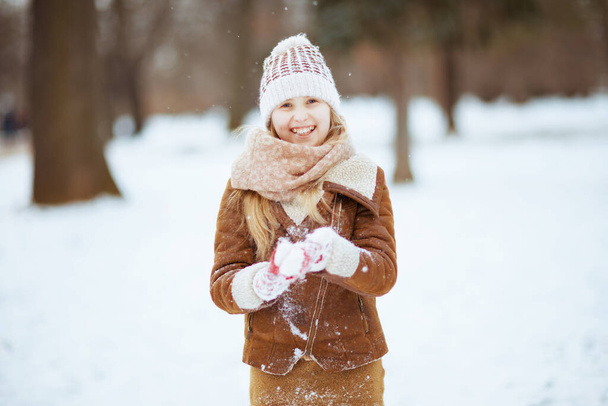 Portrait of smiling stylish girl with mittens in a knitted hat and sheepskin coat playing outside in the city park in winter. - 写真・画像