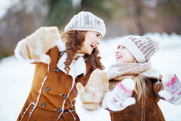 smiling elegant mother and daughter in a knitted hats and sheepskin coats outdoors in the city park in winter in a knitted hat and sheepskin coat showing snowy mittens. - Foto, Bild
