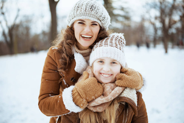 Portrait of happy elegant mother and daughter in a knitted hats and sheepskin coats with mittens in a knitted hat and sheepskin coat outdoors in the city park in winter. - Photo, Image