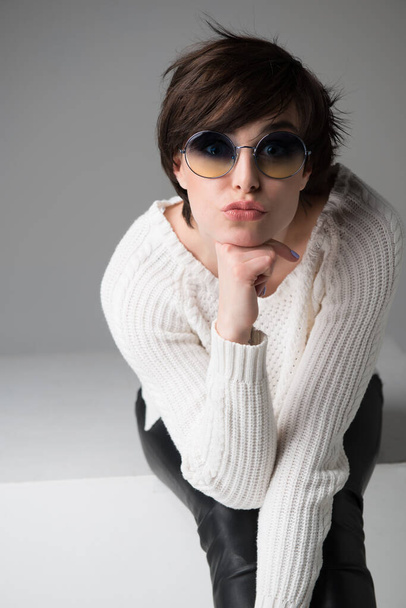 jonge vrouw met zonnebril zitten op witte kubus in studio geïsoleerd over grijze achtergrond, kijken naar camera.  - Foto, afbeelding