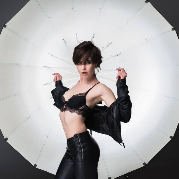 Beautiful young woman with short dark hair posing at studio in leather clothes over lightning equipment . Pretty brunette girl, studio portrait. - Fotografie, Obrázek