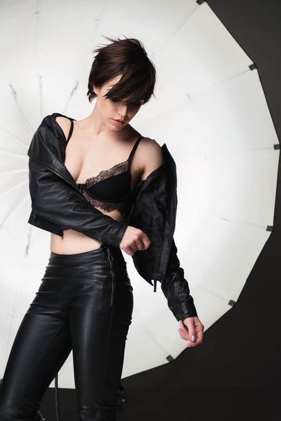 Beautiful young woman with short dark hair posing at studio in leather clothes over lightning equipment . Pretty brunette girl, studio portrait. - Photo, image