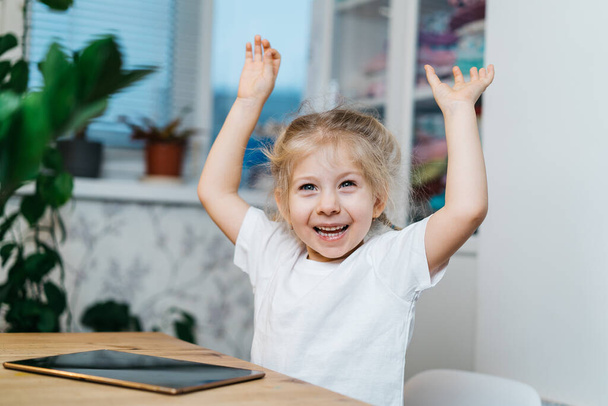 una niña está sentada en una mesa con una tableta con las manos levantadas en el aire sonriente y feliz, experimentando felicidad - Foto, imagen