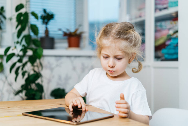 een klein meisje zit aan een tafel met een tablet met haar handen omhoog in de lucht glimlachend en gelukkig, geluk ervarend - Foto, afbeelding