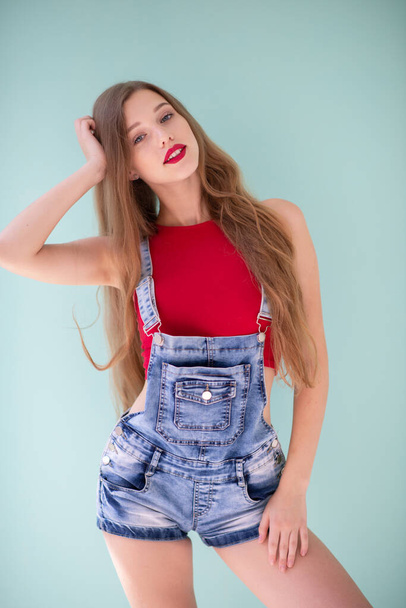 Beautiful young blond woman wearing blue jeans and red top posing indoor over light green background.  - 写真・画像